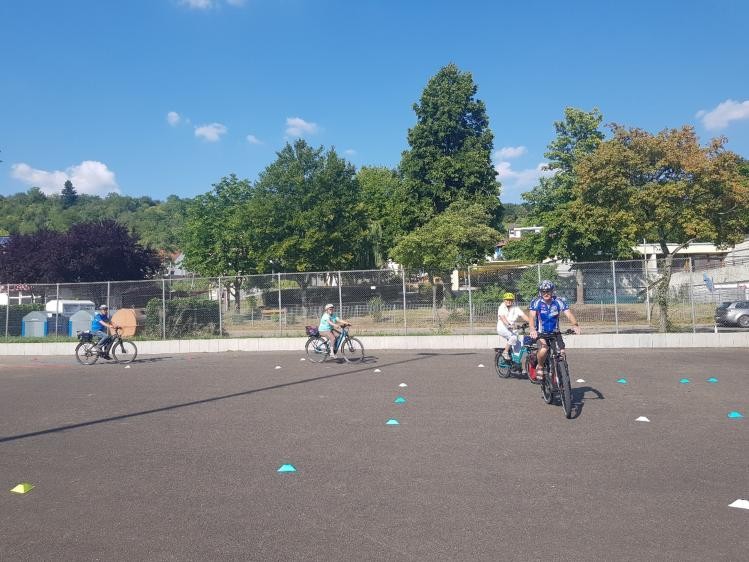 Mehrere Personen üben bei sonnigem Wetter auf einem asphaltierten Platz das Radfahren. Auf dem Boden sind farbige Markierungshütchen verteilt, die einen Übungsparcours kennzeichnen. Im Vordergrund fährt ein Mann mit Helm und Trikot auf einem Fahrrad, gefolgt von weiteren Teilnehmenden mit Helmen, darunter ein Kind. Im Hintergrund sind ein Maschendrahtzaun, Bäume, Wohnhäuser und ein blauer Himmel mit einzelnen Wolken zu sehen. Die Szene zeigt ein Fahrsicherheitstraining beziehungsweise einen Übungskurs für Radfahrende.